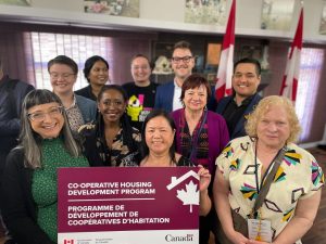 A diverse group of ten people stand together holding a sign reading "Co-operative Housing Development Program"