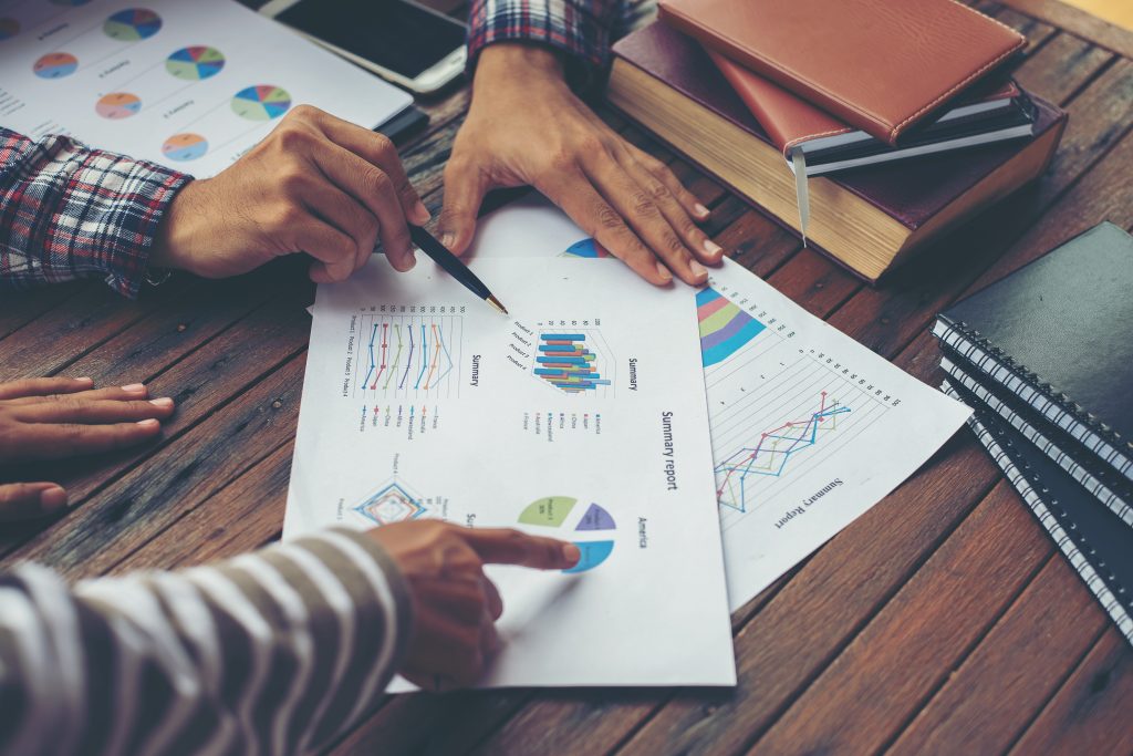 Two people's hands pointing to papers with a collection of graphs, on a table surrounded by books.